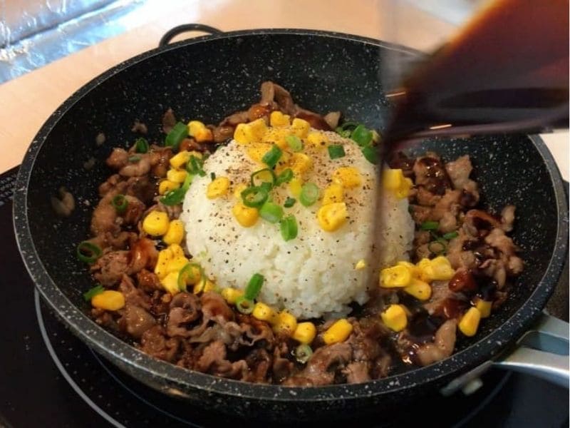 フライパンdeビーフペッパーライスにヨシダソースを回しかける調理途中の写真。ご飯、牛肉、コーン、小ネギを入れて強火で仕上げる工程。