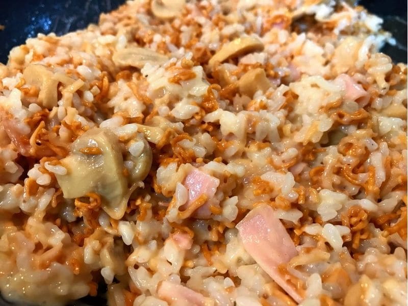 フライパンでチキンラーメンとごはんをカルボナーラソース、ハム、マッシュルームと混ぜている調理途中の写真