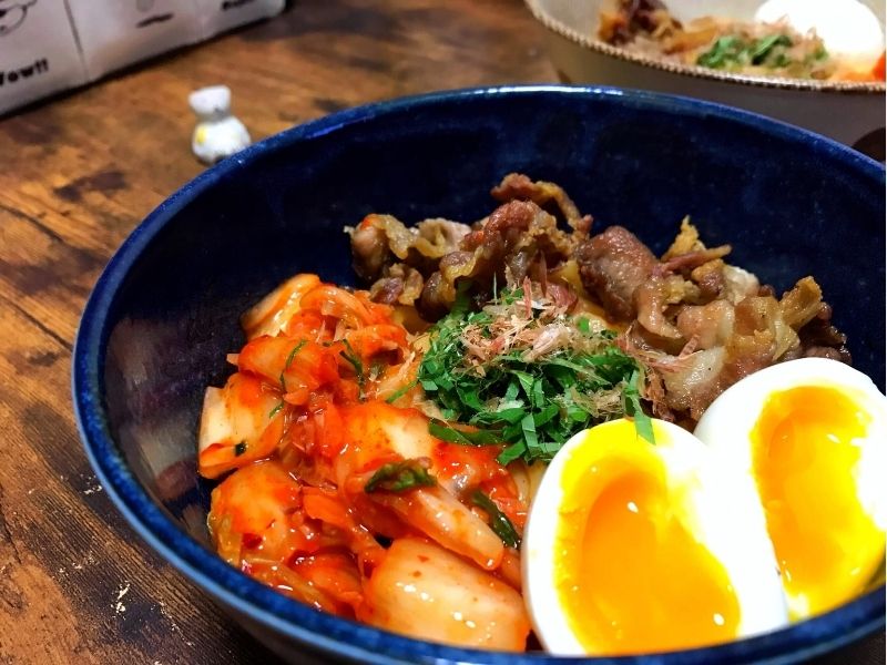 青い丼に豚こま肉、キムチ、大葉、半熟卵を盛り付けたサムギョプサル風焼肉うどん