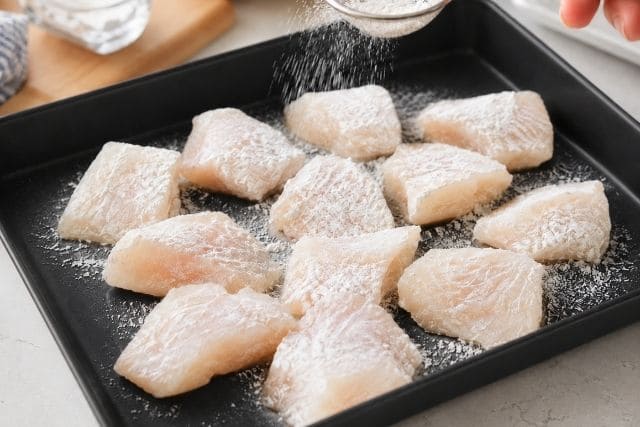 黒いトレーに並べた鱈の切り身へ片栗粉を薄くまぶしている様子