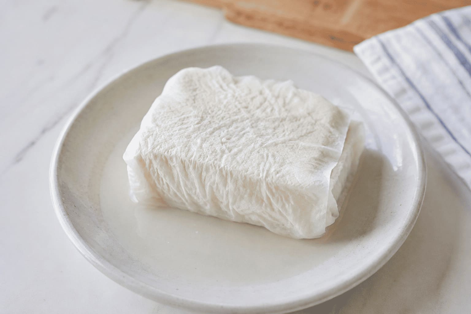 キッチンペーパーで包んだ木綿豆腐を皿にのせて水切りしている豆腐ステーキ用の下ごしらえ画像